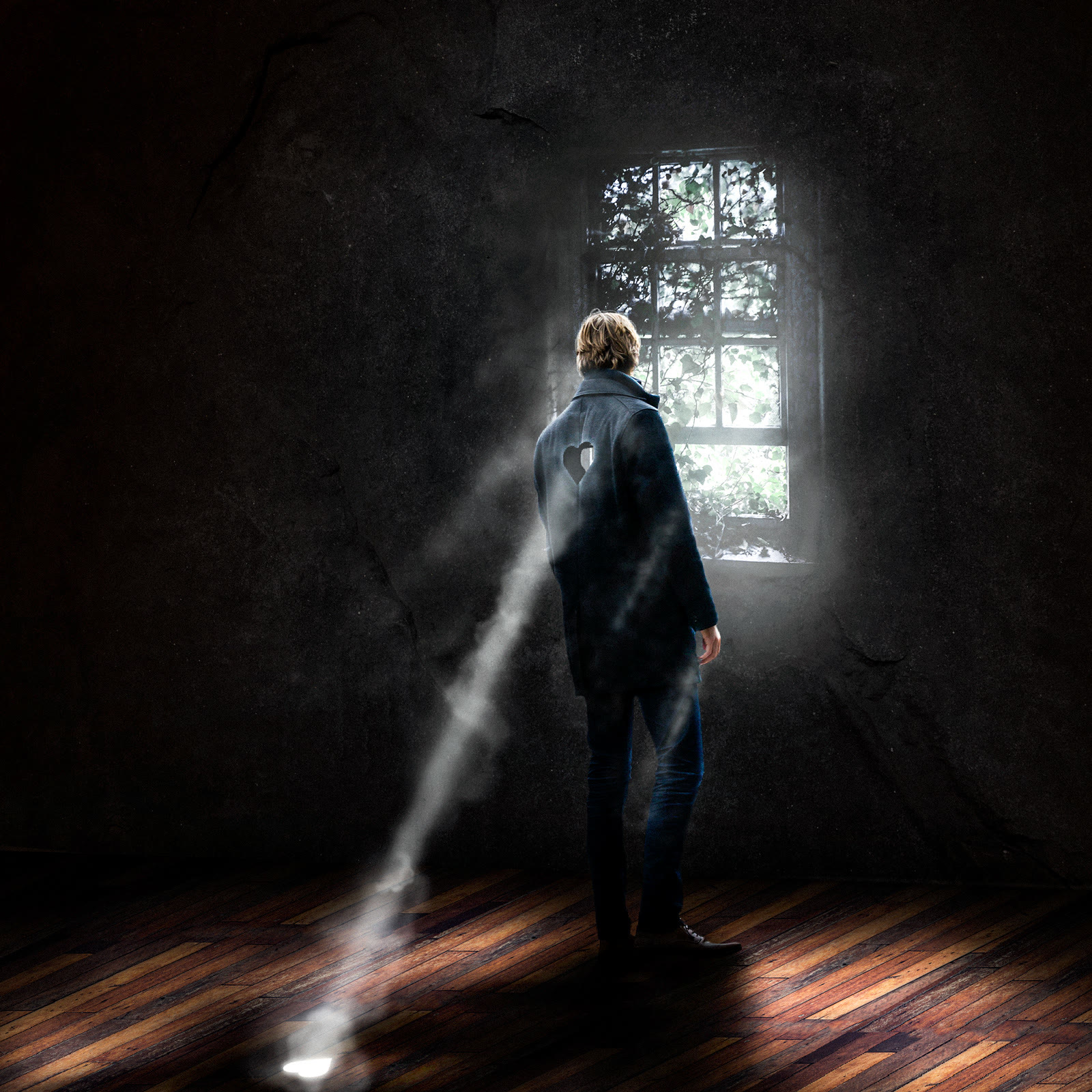 A man in a dark room facing a window, with a beam of light on the floor